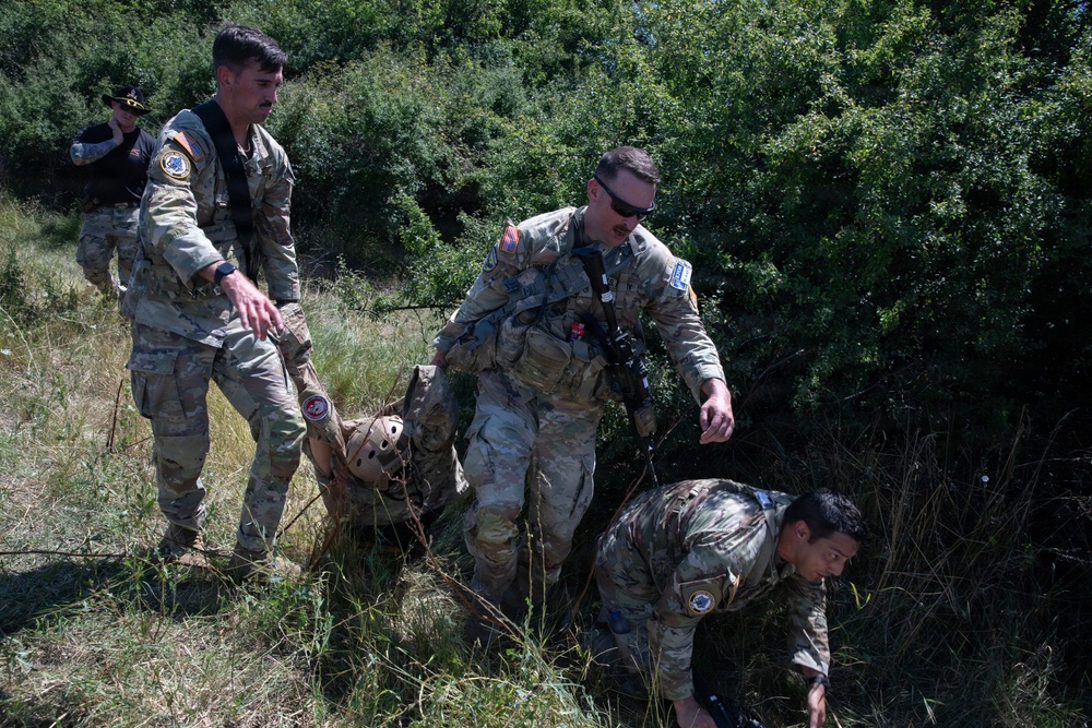Several military personnel in camouflage uniforms are conducting what appears to be a casualty evacuation drill or rescue exercise in an outdoor, vegetated terrain. Two soldiers are carrying or dragging a third person wearing a tactical helmet through brush and tall grass. A fourth individual in the background appears to be observing or documenting the exercise.