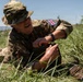 A British military soldier is lying prone in a grassy field, working with what appears to be wire or cable coiled on the ground. The soldier is wearing MultiCam camouflage uniform with a Union Jack (UK flag) patch on the shoulder and a boonie hat. The setting appears to be an outdoor training exercise or field operation.