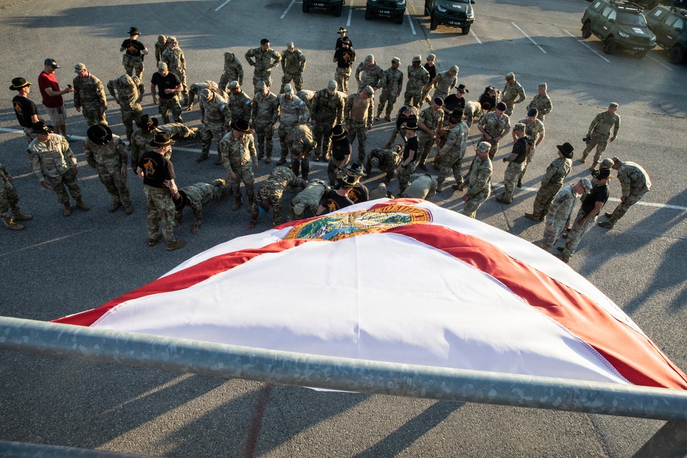 A large group of military personnel in camouflage uniforms are gathered in a parking lot around a large Florida state flag laid on the ground. Some individuals appear to be performing push-ups or physical exercises near the flag, while others stand in a circle observing. The scene appears to be a military ceremony or physical training event, photographed from an elevated angle.