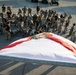 A large group of military personnel in camouflage uniforms are gathered in a parking lot around a large Florida state flag laid on the ground. Some individuals appear to be performing push-ups or physical exercises near the flag, while others stand in a circle observing. The scene appears to be a military ceremony or physical training event, photographed from an elevated angle.