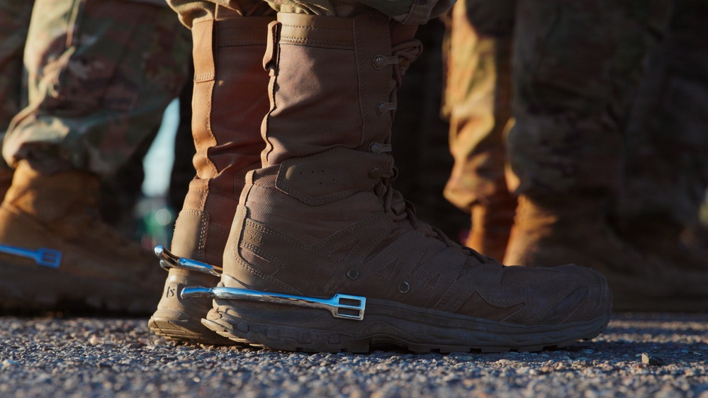 Close-up photograph of military personnel's lower legs and boots, focused on a tan/coyote brown tactical combat boot. A shiny chrome or polished metal spur is attached to the heel of the foreground boot, suggesting a military spurs ceremony or award. Multiple individuals in camouflage uniforms are visible in the background.