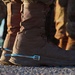 Close-up photograph of military personnel's lower legs and boots, focused on a tan/coyote brown tactical combat boot. A shiny chrome or polished metal spur is attached to the heel of the foreground boot, suggesting a military spurs ceremony or award. Multiple individuals in camouflage uniforms are visible in the background.
