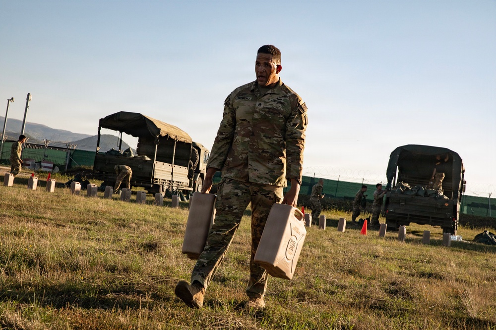 A U.S. Army soldier in MultiCam/OCP camouflage uniform is carrying two tan military-issue jerry cans across a grassy field at what appears to be a forward operating base or training area. Multiple military trucks with canvas covers are visible in the background, along with other soldiers conducting logistics or supply operations. The scene suggests a field exercise or operational resupply activity during early morning or late afternoon light.