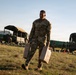 A U.S. Army soldier in MultiCam/OCP camouflage uniform is carrying two tan military-issue jerry cans across a grassy field at what appears to be a forward operating base or training area. Multiple military trucks with canvas covers are visible in the background, along with other soldiers conducting logistics or supply operations. The scene suggests a field exercise or operational resupply activity during early morning or late afternoon light.