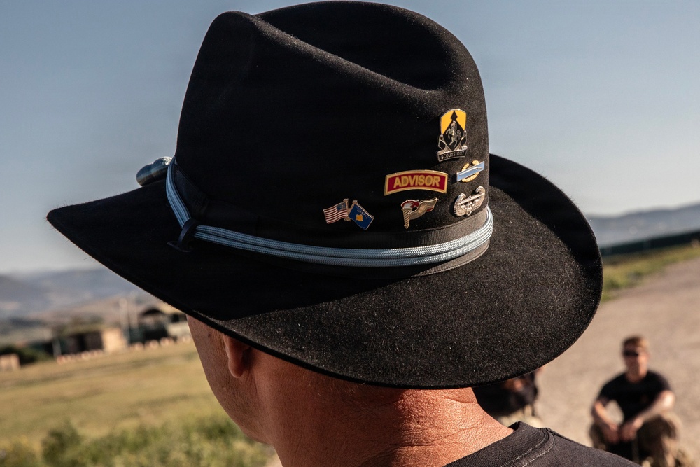 A close-up photograph of a black felt cowboy/campaign-style hat adorned with multiple military and commemorative pins on its hatband. The hat is worn by a person whose face is not visible, photographed from behind. The setting appears to be an outdoor military or training environment with arid terrain visible in the background.