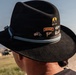 A close-up photograph of a black felt cowboy/campaign-style hat adorned with multiple military and commemorative pins on its hatband. The hat is worn by a person whose face is not visible, photographed from behind. The setting appears to be an outdoor military or training environment with arid terrain visible in the background.