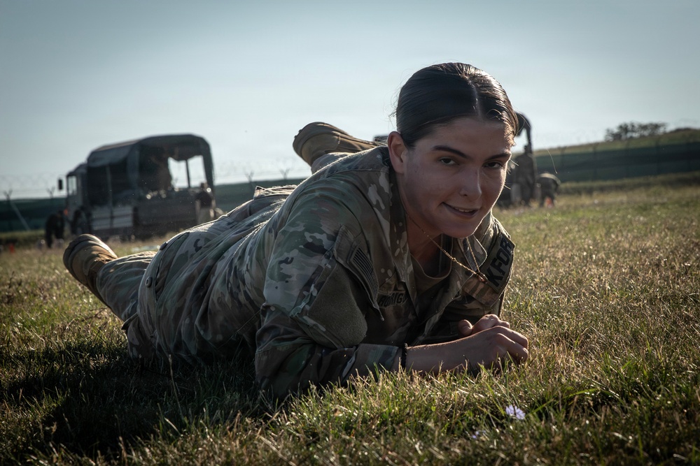 A female soldier in U.S. Army MultiCam/OCP camouflage uniform is performing a low crawl on a grass field, smiling toward the camera. She appears to be engaged in a physical training or field exercise. Military vehicles and other personnel are visible in the background.