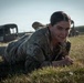 A female soldier in U.S. Army MultiCam/OCP camouflage uniform is performing a low crawl on a grass field, smiling toward the camera. She appears to be engaged in a physical training or field exercise. Military vehicles and other personnel are visible in the background.