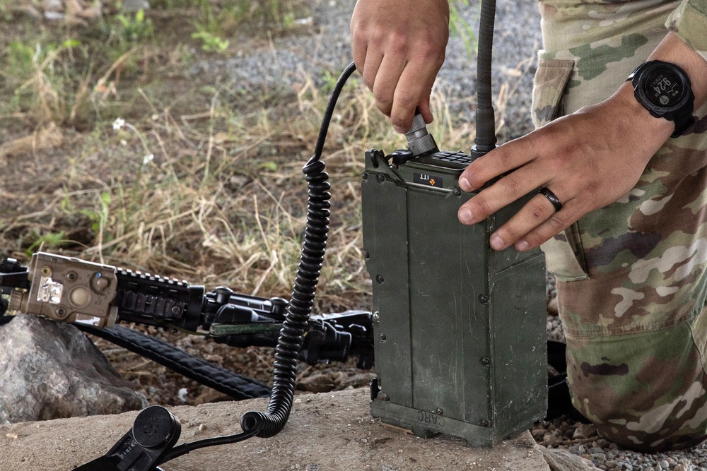 A soldier in MultiCam/OCP camouflage uniform is operating a military field radio, identifiable by the label 'ARC' on its olive drab casing. The individual is connecting or adjusting a coiled cable to the radio unit, which has an antenna attached. A military rifle with accessories is visible on the ground nearby.