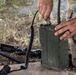 A soldier in MultiCam/OCP camouflage uniform is operating a military field radio, identifiable by the label 'ARC' on its olive drab casing. The individual is connecting or adjusting a coiled cable to the radio unit, which has an antenna attached. A military rifle with accessories is visible on the ground nearby.