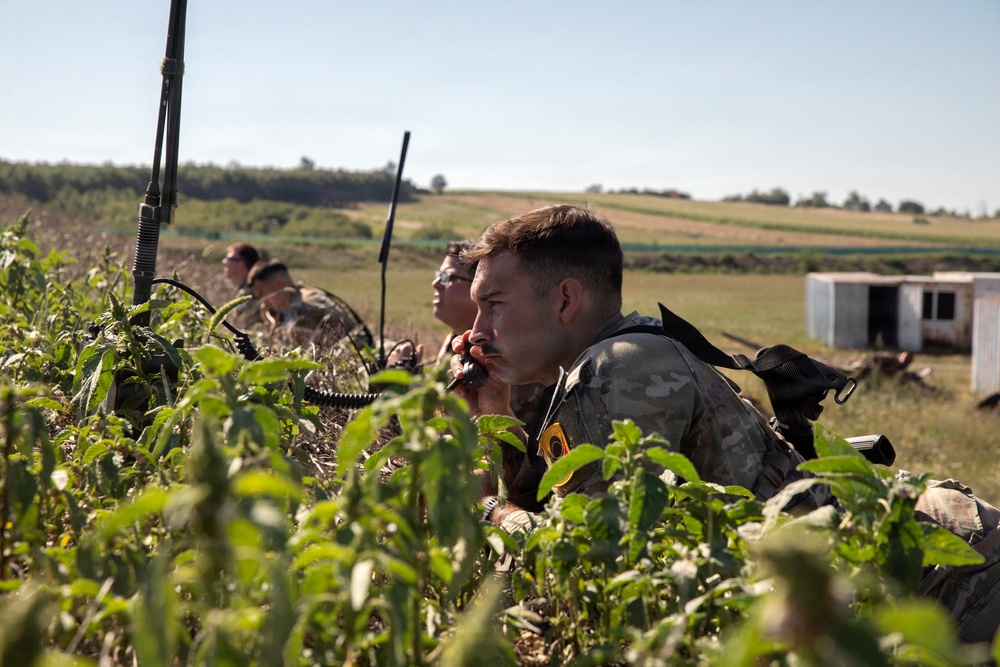 Three military personnel in camouflage uniforms are positioned low in vegetation, appearing to conduct a field observation or reconnaissance exercise. The foreground soldier is holding a radio handset to his mouth while wearing a distinctive yellow patch on his uniform. The setting appears to be a rural training area with open fields and a small structure visible in the background.