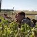Three military personnel in camouflage uniforms are positioned low in vegetation, appearing to conduct a field observation or reconnaissance exercise. The foreground soldier is holding a radio handset to his mouth while wearing a distinctive yellow patch on his uniform. The setting appears to be a rural training area with open fields and a small structure visible in the background.