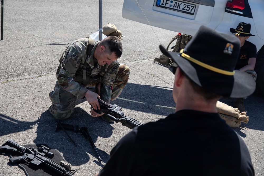 A U.S. Army soldier in MultiCam camouflage uniform with a 'KFOR' patch is kneeling on pavement, handling and demonstrating a weapon, likely during a public display or demonstration event. A civilian or off-duty personnel wearing a black cavalry-style hat with gold trim and a black shirt observes from the foreground. Additional weapons and equipment are visible on the ground nearby.