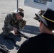 A U.S. Army soldier in MultiCam camouflage uniform with a 'KFOR' patch is kneeling on pavement, handling and demonstrating a weapon, likely during a public display or demonstration event. A civilian or off-duty personnel wearing a black cavalry-style hat with gold trim and a black shirt observes from the foreground. Additional weapons and equipment are visible on the ground nearby.