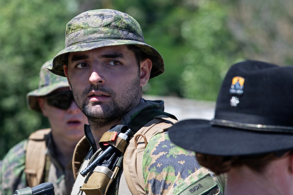 The image shows three individuals in military or tactical gear in an outdoor setting with green vegetation in the background. The central figure, wearing a camouflage boonie hat and tactical vest with equipment, appears to be the primary subject in sharp focus. A name tape reading 'IBISHI' is visible on one of the individuals.
