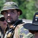 The image shows three individuals in military or tactical gear in an outdoor setting with green vegetation in the background. The central figure, wearing a camouflage boonie hat and tactical vest with equipment, appears to be the primary subject in sharp focus. A name tape reading 'IBISHI' is visible on one of the individuals.