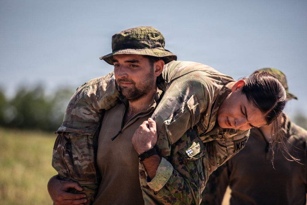 Two military personnel in camouflage uniforms are engaged in what appears to be a casualty carry drill or combat training exercise. A female soldier wearing U.S. Army-style OCP uniform is performing a fireman's carry or buddy carry on a male soldier wearing a camouflage boonie hat. The setting appears to be an outdoor field training environment with trees visible in the background.