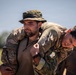 Two military personnel in camouflage uniforms are engaged in what appears to be a casualty carry drill or combat training exercise. A female soldier wearing U.S. Army-style OCP uniform is performing a fireman's carry or buddy carry on a male soldier wearing a camouflage boonie hat. The setting appears to be an outdoor field training environment with trees visible in the background.