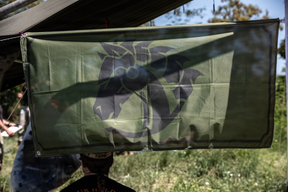 A green flag or banner featuring a dark stylized emblem — appearing to depict a horse or dragon-like creature with a circular element — is suspended under a canopy or awning in an outdoor setting. A person wearing a cap with the name 'PAYNE' visible is standing beneath the flag. The setting appears to be a field or outdoor gathering, possibly military or tactical in nature.