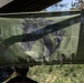 A green flag or banner featuring a dark stylized emblem — appearing to depict a horse or dragon-like creature with a circular element — is suspended under a canopy or awning in an outdoor setting. A person wearing a cap with the name 'PAYNE' visible is standing beneath the flag. The setting appears to be a field or outdoor gathering, possibly military or tactical in nature.