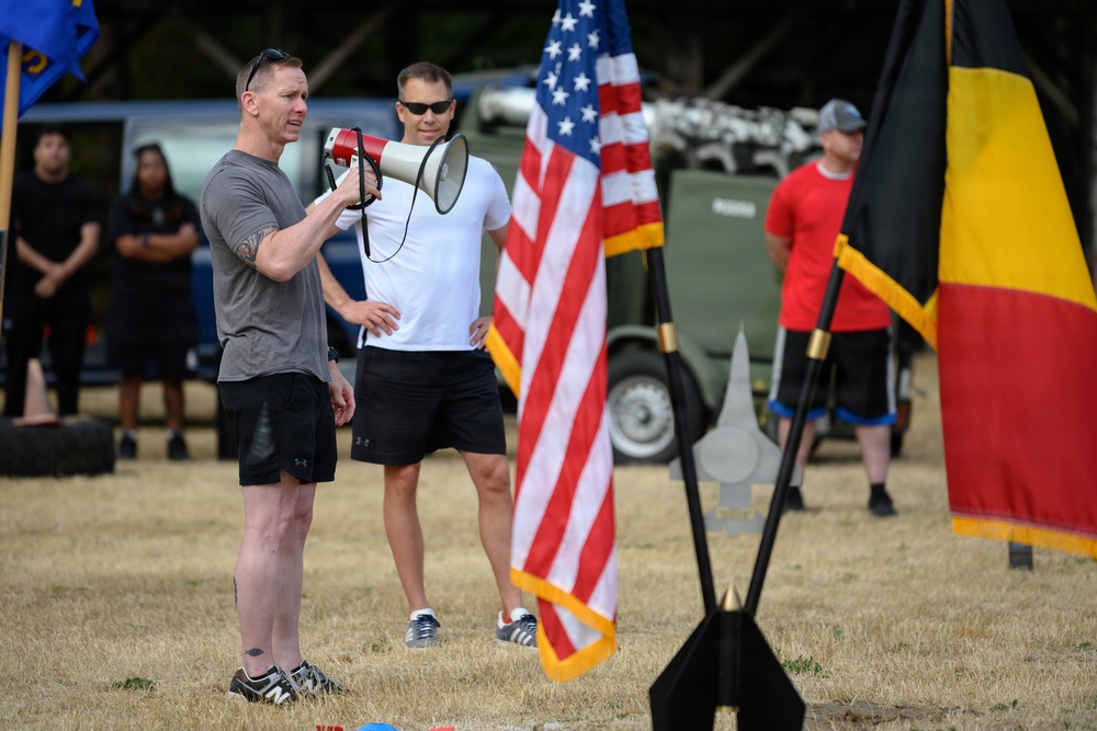 An outdoor military or athletic event featuring two men in athletic wear, one speaking through a red and white megaphone. An American flag and what appears to be a Belgian flag (black, yellow, red vertical stripes) are prominently displayed. Several other participants are visible in the background near military-style vehicles.