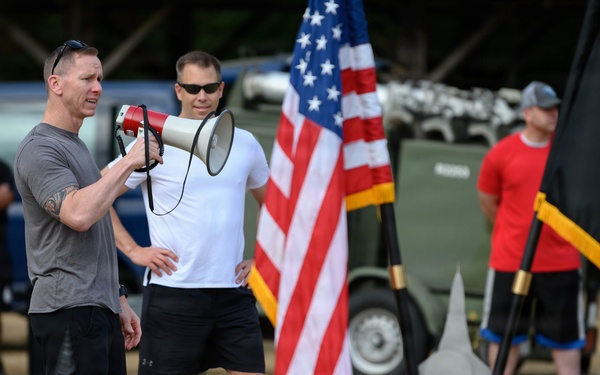 An outdoor military or athletic event featuring two men in athletic wear, one speaking through a red and white megaphone. An American flag and what appears to be a Belgian flag (black, yellow, red vertical stripes) are prominently displayed. Several other participants are visible in the background near military-style vehicles.