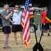 An outdoor military or athletic event featuring two men in athletic wear, one speaking through a red and white megaphone. An American flag and what appears to be a Belgian flag (black, yellow, red vertical stripes) are prominently displayed. Several other participants are visible in the background near military-style vehicles.