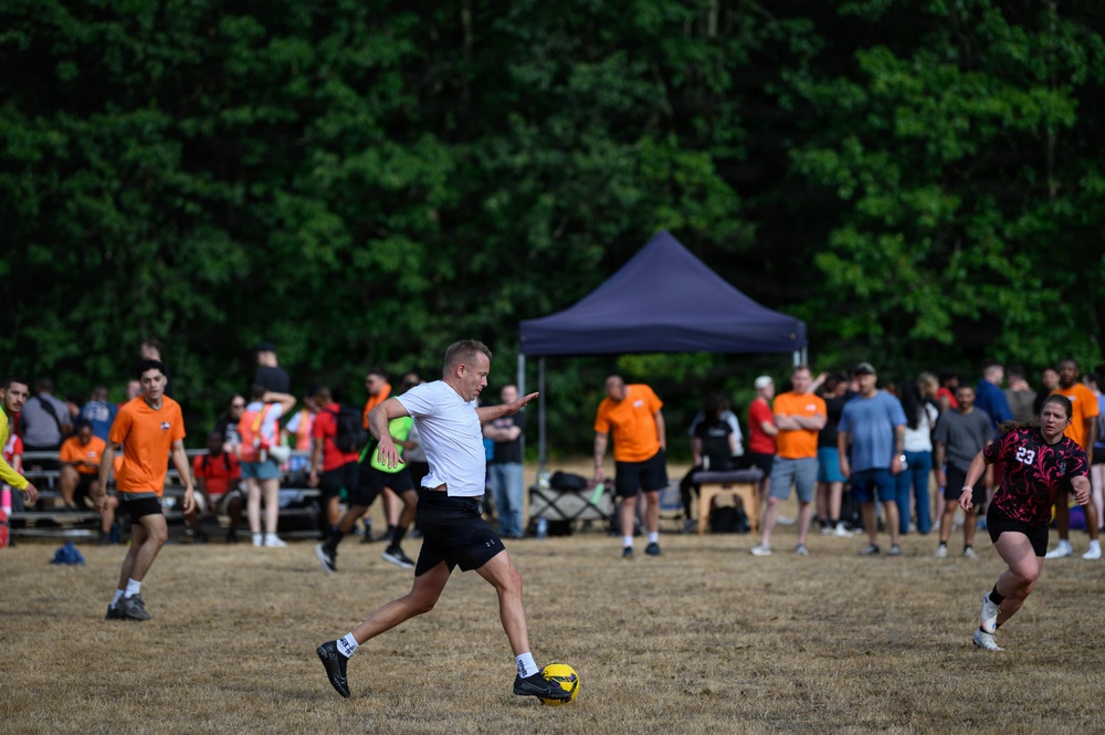 An outdoor soccer match is taking place on a dry grass field, with a player in a white shirt about to kick a yellow soccer ball. Multiple teams in different colored jerseys (orange, red, black/pink) are visible, along with a large crowd of spectators gathered near a dark blue canopy tent. The setting appears to be a casual or recreational tournament in a park or open field surrounded by dense green trees.