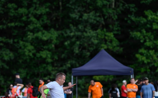 An outdoor soccer match is taking place on a dry grass field, with a player in a white shirt about to kick a yellow soccer ball. Multiple teams in different colored jerseys (orange, red, black/pink) are visible, along with a large crowd of spectators gathered near a dark blue canopy tent. The setting appears to be a casual or recreational tournament in a park or open field surrounded by dense green trees.