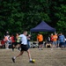 An outdoor soccer match is taking place on a dry grass field, with a player in a white shirt about to kick a yellow soccer ball. Multiple teams in different colored jerseys (orange, red, black/pink) are visible, along with a large crowd of spectators gathered near a dark blue canopy tent. The setting appears to be a casual or recreational tournament in a park or open field surrounded by dense green trees.