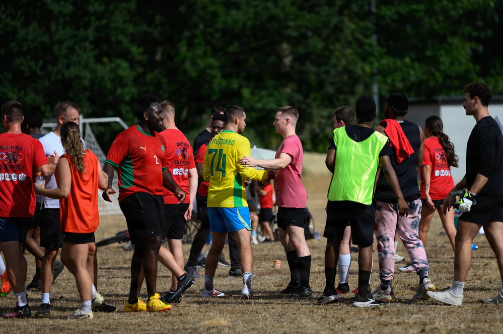 A group of players gathered on a grass football pitch following what appears to be a recreational or community football match. Players are wearing various team kits including red jerseys labeled 'MUNSS OLYMPICS' and a yellow/green Brazil-style kit with number 74 ('TOSDINHO'). Two players in the center appear to be shaking hands in a post-match gesture.