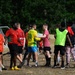 A group of players gathered on a grass football pitch following what appears to be a recreational or community football match. Players are wearing various team kits including red jerseys labeled 'MUNSS OLYMPICS' and a yellow/green Brazil-style kit with number 74 ('TOSDINHO'). Two players in the center appear to be shaking hands in a post-match gesture.