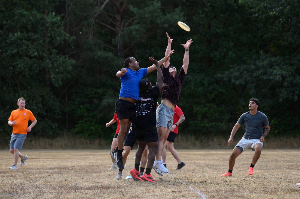 A group of players are engaged in a competitive game of Ultimate Frisbee on a dry grass field. The central action shows multiple players jumping and reaching for a yellow frisbee in mid-air, with one player in a blue shirt contesting against a player in a dark maroon shirt. Additional players are visible in the background, including one in an orange shirt acting as a referee or observer.