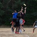 A group of players are engaged in a competitive game of Ultimate Frisbee on a dry grass field. The central action shows multiple players jumping and reaching for a yellow frisbee in mid-air, with one player in a blue shirt contesting against a player in a dark maroon shirt. Additional players are visible in the background, including one in an orange shirt acting as a referee or observer.
