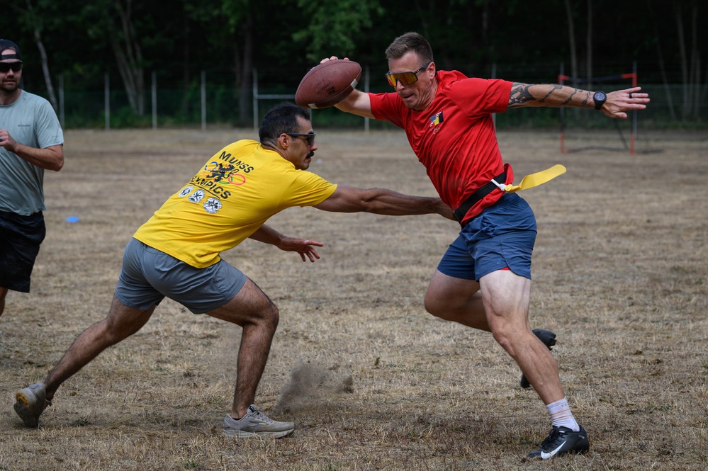 Two men are playing flag football on a dry grass field. The player in a red shirt is holding a football aloft while the defender in a yellow shirt attempts to pull his flag. A third person in a gray shirt is visible in the background.