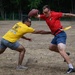 Two men are playing flag football on a dry grass field. The player in a red shirt is holding a football aloft while the defender in a yellow shirt attempts to pull his flag. A third person in a gray shirt is visible in the background.
