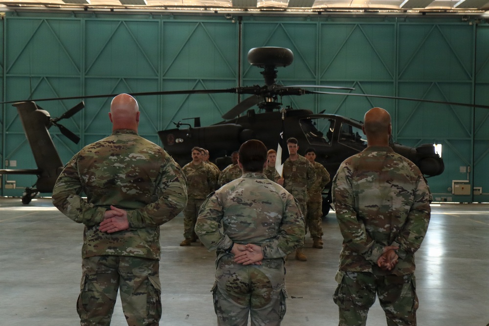 Military personnel in U.S. Army MultiCam/OCP camouflage uniforms are gathered inside a large aircraft hangar. Three soldiers stand with hands clasped behind their backs facing a group of additional soldiers, with an AH-64 Apache attack helicopter visible in the background. The setting appears to be a formal briefing or formation inside the hangar.