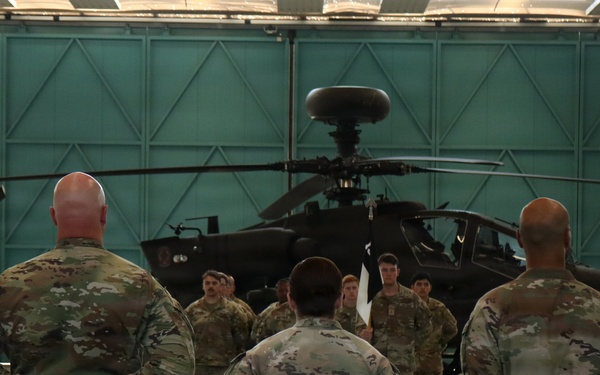 Military personnel in U.S. Army MultiCam/OCP camouflage uniforms are gathered inside a large aircraft hangar. Three soldiers stand with hands clasped behind their backs facing a group of additional soldiers, with an AH-64 Apache attack helicopter visible in the background. The setting appears to be a formal briefing or formation inside the hangar.