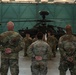 Military personnel in U.S. Army MultiCam/OCP camouflage uniforms are gathered inside a large aircraft hangar. Three soldiers stand with hands clasped behind their backs facing a group of additional soldiers, with an AH-64 Apache attack helicopter visible in the background. The setting appears to be a formal briefing or formation inside the hangar.