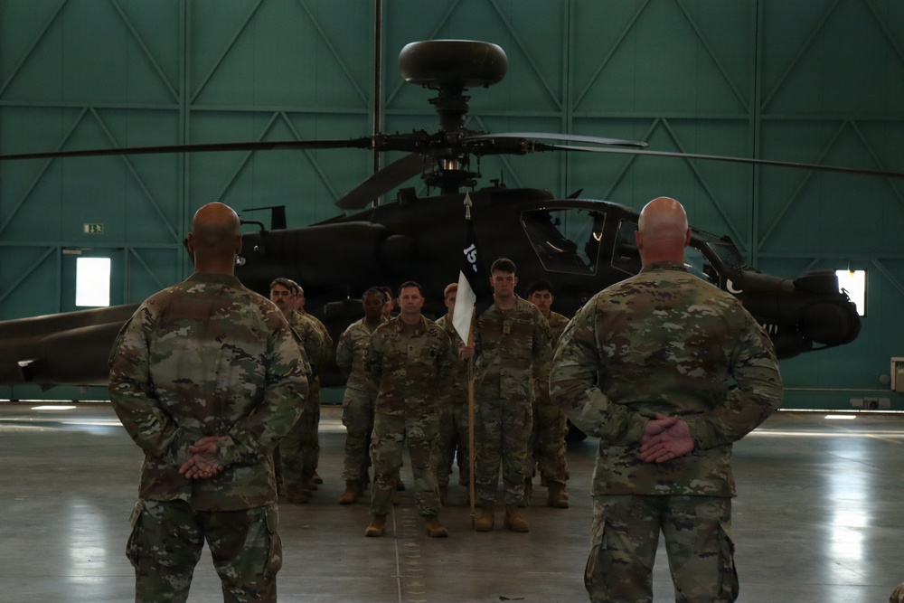 A military formation ceremony is taking place inside a large aircraft hangar, with soldiers in camouflage uniforms standing at attention before two senior personnel. A soldier in the center of the formation holds a unit guidon (flag). An AH-64 Apache attack helicopter is prominently visible in the background.