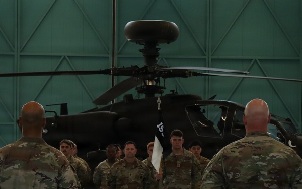A military formation ceremony is taking place inside a large aircraft hangar, with soldiers in camouflage uniforms standing at attention before two senior personnel. A soldier in the center of the formation holds a unit guidon (flag). An AH-64 Apache attack helicopter is prominently visible in the background.