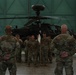 A military formation ceremony is taking place inside a large aircraft hangar, with soldiers in camouflage uniforms standing at attention before two senior personnel. A soldier in the center of the formation holds a unit guidon (flag). An AH-64 Apache attack helicopter is prominently visible in the background.