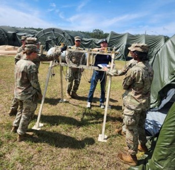 A group of military personnel in camouflage uniforms are gathered around a white metal frame structure on a grassy field, appearing to be engaged in a training or equipment assembly exercise. One civilian in jeans and a dark shirt is present among the soldiers, possibly serving as an instructor or technical advisor. The setting appears to be a military field camp, evidenced by multiple large green military tents visible in the background.