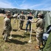 A group of military personnel in camouflage uniforms are gathered around a white metal frame structure on a grassy field, appearing to be engaged in a training or equipment assembly exercise. One civilian in jeans and a dark shirt is present among the soldiers, possibly serving as an instructor or technical advisor. The setting appears to be a military field camp, evidenced by multiple large green military tents visible in the background.