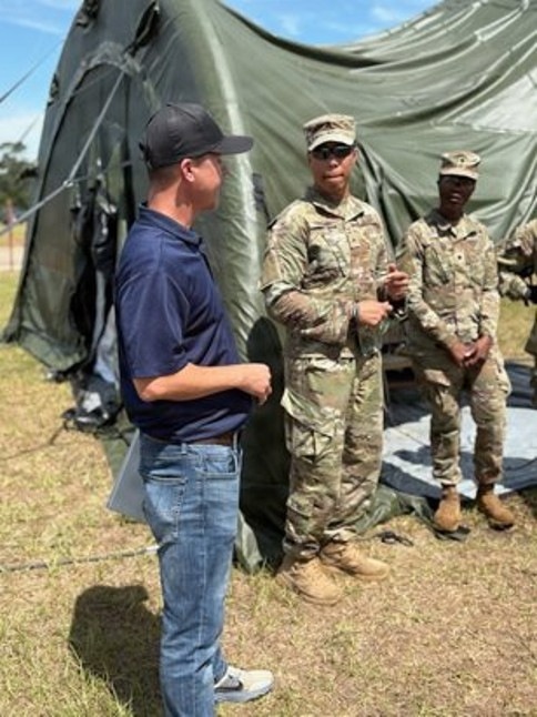A civilian in casual attire (navy polo shirt, jeans, baseball cap) is engaged in conversation with two soldiers wearing US Army OCP (Operational Camouflage Pattern) uniforms. The interaction takes place outdoors on a grassy field in front of a large military-style olive drab tent. The setting appears to be a field training exercise or military installation.