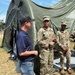 A civilian in casual attire (navy polo shirt, jeans, baseball cap) is engaged in conversation with two soldiers wearing US Army OCP (Operational Camouflage Pattern) uniforms. The interaction takes place outdoors on a grassy field in front of a large military-style olive drab tent. The setting appears to be a field training exercise or military installation.