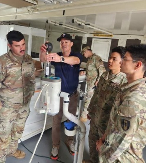 A civilian instructor or technician in a navy blue polo shirt and baseball cap is demonstrating water purification or filtration equipment to a group of military personnel in OCP (Operational Camouflage Pattern) uniforms. The setting appears to be inside a mobile training unit or containerized facility. The instructor is holding a pressure gauge while explaining the equipment's components to attentive soldiers.