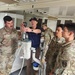 A civilian instructor or technician in a navy blue polo shirt and baseball cap is demonstrating water purification or filtration equipment to a group of military personnel in OCP (Operational Camouflage Pattern) uniforms. The setting appears to be inside a mobile training unit or containerized facility. The instructor is holding a pressure gauge while explaining the equipment's components to attentive soldiers.