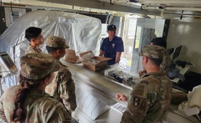 Several military personnel in OCP (Operational Camouflage Pattern) uniforms are gathered at what appears to be a supply or equipment distribution counter. A civilian or off-duty personnel in a dark polo shirt stands behind the counter, which has open boxes and equipment on it. The setting appears to be a temporary or field facility, indicated by the plastic sheeting visible in the background.
