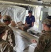 Several military personnel in OCP (Operational Camouflage Pattern) uniforms are gathered at what appears to be a supply or equipment distribution counter. A civilian or off-duty personnel in a dark polo shirt stands behind the counter, which has open boxes and equipment on it. The setting appears to be a temporary or field facility, indicated by the plastic sheeting visible in the background.